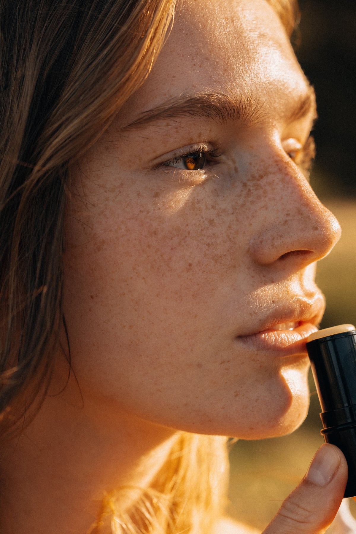 Close-up of a person applying lipstick outdoors with a blurred natural background