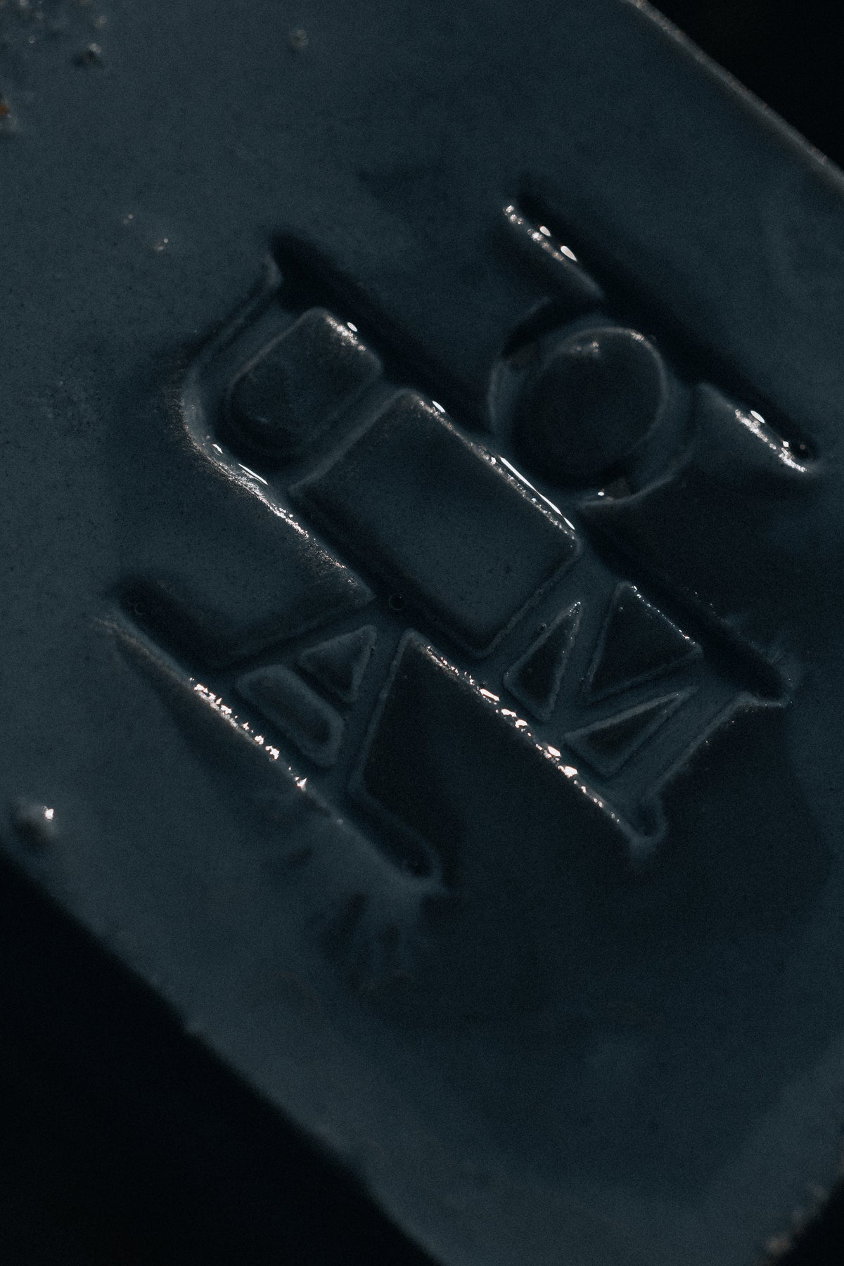 Close-up of a dark teal-blue rectangular product with an embossed stylized logo or lettering, photographed against a black background.