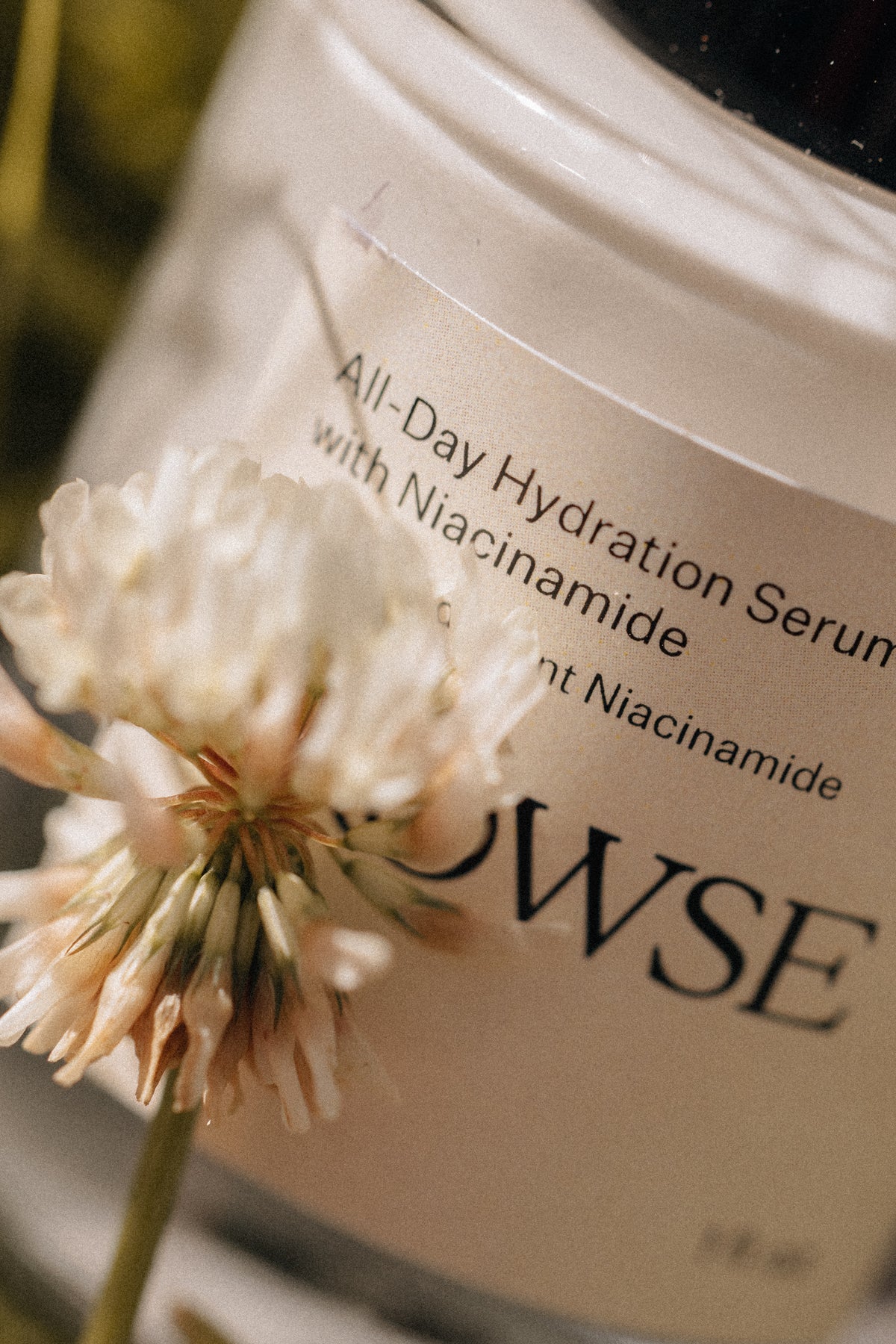 Cosmetic jar labeled 'All-Day Hydration Serum with Niacinamide' by 'LOWSE' with a white flower in front.